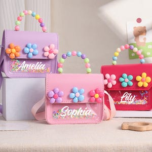 May include: Three small handbags in purple, light pink, and hot pink. Each bag features a top handle made of colorful plastic beads, three plastic flower embellishments on the flap, and a clear front pocket containing confetti with the names Amelia, Sophia, or Lily.