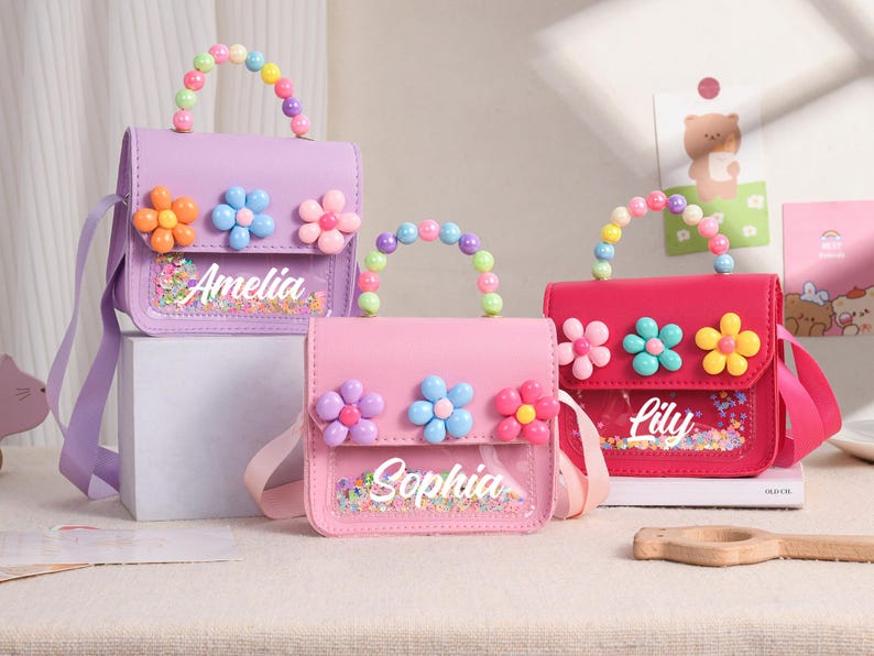 May include: Three small handbags in purple, light pink, and hot pink. Each bag features a top handle made of colorful plastic beads, three plastic flower embellishments on the flap, and a clear front pocket containing confetti with the names Amelia, Sophia, or Lily.