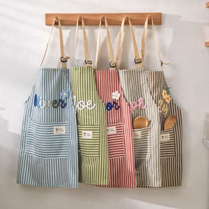 May include: Five striped aprons hanging on a wooden rack. Each apron has a different color scheme, including blue, green, red, gray, and brown. The aprons have pockets and decorative floral or text embellishments.