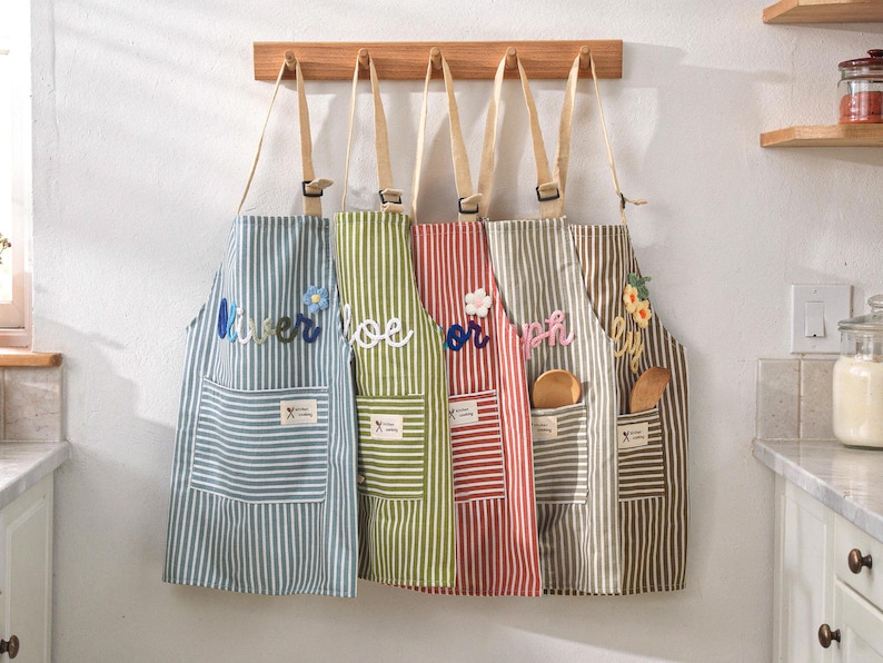 May include: Five striped aprons hanging on a wooden rack. Each apron has a different color scheme, including blue, green, red, gray, and brown. The aprons have pockets and decorative floral or text embellishments.