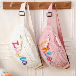 Op de afbeelding: Twee fluffy fanny packs in sherpa-stijl hangen aan een houten rek. Eén is wit met kleurrijke geborduurde tekst "Luv ba". De andere is roze met "Love" geborduurd in kleurrijk garen. Beide hebben verstelbare bandjes.
