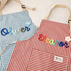 May include: Two striped aprons with personalized names. One is blue and white with the name "Oliver" in blue, green, and white yarn. The other is red and white with the name "Eleanor" in rainbow yarn. Both have pockets and adjustable straps.