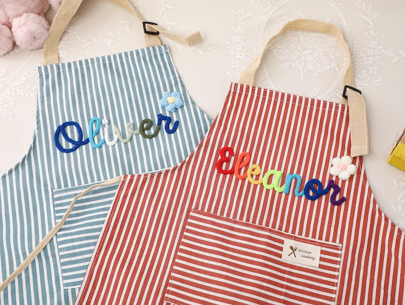 May include: Two striped aprons with personalized names. One is blue and white with the name "Oliver" in blue, green, and white yarn. The other is red and white with the name "Eleanor" in rainbow yarn. Both have pockets and adjustable straps.