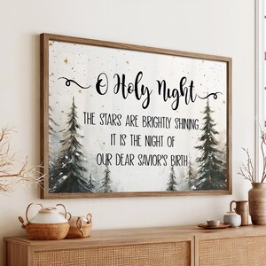 以下が含まれることがあります： 「O Holy Night」のテキストがエレガントな筆記体で書かれた額入りのウォールアートプリント。このアートワークは、常緑樹と星空のある冬の森の風景が特徴です。下のテキストは「星は明るく輝いています。それは私たちの最愛の救い主の誕生の夜です」と書かれています。