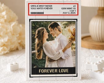 Puede incluir: Un expositor rectangular de bloques de construcción blancos con una fotografía de una pareja abrazada. La parte superior incluye el texto GREG & MISSY MOSER, SOUL MATES FOREVER, #1, GEM MINT 10, 1 OF 1, JULY 2, 2026. La parte inferior muestra FOREVER LOVE.