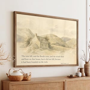 May include: A framed watercolor print featuring a quaint cottage nestled in a mountainous landscape. The artwork is in muted tones of beige, gray, and brown. Text from Matthew 7:25 is printed below the image.