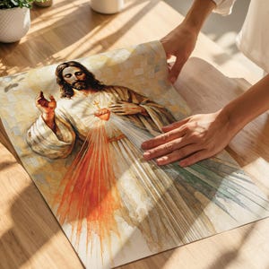 May include: A religious print depicting Jesus with a radiant heart and outstretched hand. The artwork features warm tones of beige, orange, and white, with detailed brushstrokes. The image is held by hands, suggesting it's a print or poster.