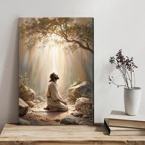 May include: A canvas print depicting a figure kneeling in prayer under a tree, bathed in sunlight. The artwork features a serene scene with soft colors, rocks, and foliage, evoking a sense of peace and spirituality. The figure is wearing a light-colored robe.
