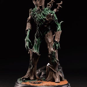 Op de afbeelding: Gedetailleerd beeldje van een boomachtig wezen met een mensachtige vorm. De figuur heeft een bruine, getextureerde, schorsachtige buitenkant met groen, mosachtig gebladerte. Het staat op een cirkelvormige basis.