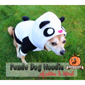 May include: A small dog wearing a white and black panda hooded sweatshirt. The sweatshirt has a black eye patch and a pink nose. The dog is sitting on green grass. The text "Panda Dog Hoodie pdf pattern & tutorial" is at the bottom of the image.