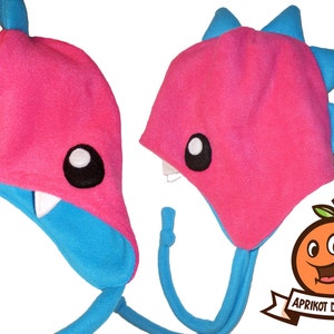 May include: A pair of handmade, plush hats in a whimsical monster design. The hats are primarily pink with blue accents, featuring large white-rimmed eyes, white teeth, and a blue fin or spike detail. The hats have long, blue dangling ties.