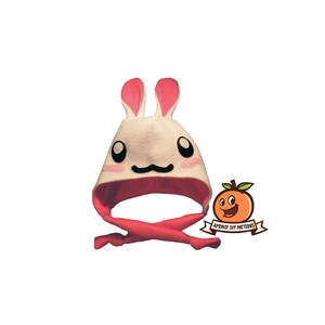 May include: A white bunny hat with pink ears and a pink fleece lining. The hat has a tie at the bottom and a cute cartoon face.
