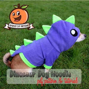Puede incluir: Una sudadera con capucha para perro de color morado y verde con forma de dinosaurio, con capucha y cola. La sudadera tiene una cara linda con ojos grandes. El texto "Dinosaur Dog Hoodie pdf pattern & tutorial" está en la parte inferior de la imagen.