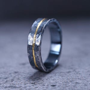 Puede incluir: Un anillo de metal gris oscuro con un acabado martillado y texturizado en toda su superficie exterior. Una incrustaci&oacute;n vertical fina, lisa y de color dorado recorre el centro de la banda, creando un contraste marcado con el metal oscuro.