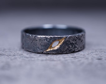 Op de afbeelding: Een close-up van een getextureerde, donkergrijze ring met een goudkleurige inleg. De ring heeft een ruwe, organische uitstraling, met een klein, ovaalvormig goudkleurig accent. De ring staat tegen een vage grijze achtergrond.