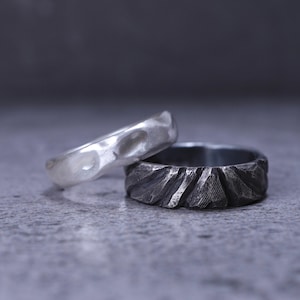May include: Two rings are displayed together. One ring is bright silver with a smooth, undulating surface featuring rounded indentations. The second ring is dark, oxidized silver with a rugged, textured surface composed of sharp, angular facets.