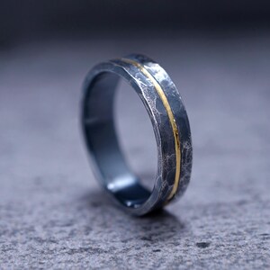 Puede incluir: Un anillo gris oscuro con un acabado martillado y texturizado, con una banda fina y lisa de color dorado que recorre horizontalmente el centro. El anillo tiene un perfil redondeado y un brillo met&aacute;lico, mostrando el contraste entre el exterior oscuro y rugoso y la incrustaci&oacute;n brillante y pulida de color dorado.
