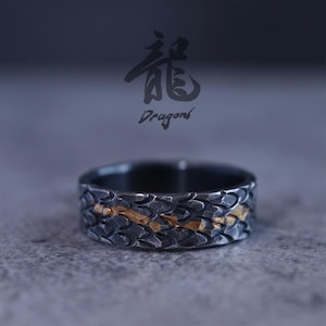 Peut inclure: Une bague gris foncé avec un motif texturé en forme d'écailles et un accent doré. Le mot "Dragons" est écrit au-dessus de la bague dans une police stylisée.