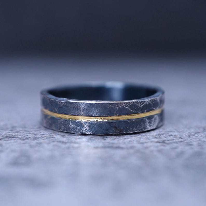 Puede incluir: Un anillo gris oscuro con un acabado texturizado y martillado, con una fina banda horizontal de color dorado en el centro. La superficie met&aacute;lica parece oxidada, creando un contraste entre el exterior oscuro y rugoso y la incrustaci&oacute;n dorada lisa.