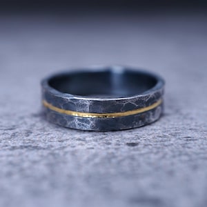 Puede incluir: Un anillo gris oscuro con un acabado martillado y texturizado, que presenta una fina banda horizontal de color dorado en el centro. El anillo tiene un perfil plano y un aspecto mate, destacando el contraste entre el metal oscuro y la incrustaci&oacute;n dorada.