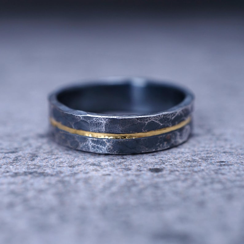 Puede incluir: Un anillo gris oscuro con un acabado martillado y texturizado, que presenta una fina banda horizontal de color dorado en el centro. El anillo tiene un perfil plano y un aspecto mate, destacando el contraste entre el metal oscuro y la incrustaci&oacute;n dorada.