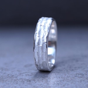 Puede incluir: Un anillo de tono plateado que presenta una superficie exterior texturizada y martillada con un patrón repetitivo de hendiduras verticales. La banda tiene un interior pulido y reflectante con un acabado metálico que capta la luz a lo largo de su borde exterior irregular.