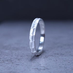 Puede incluir: Un anillo de plata con un acabado martillado y texturizado en la banda exterior. La superficie metálica presenta hendiduras irregulares y reflectantes que crean una apariencia facetada. El anillo está colocado en posición vertical.