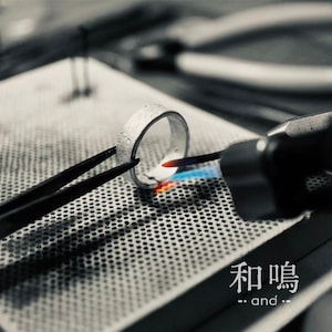 Puede incluir: Un anillo de plata sostenido por pinzas de metal negro mientras un soplete manual aplica una llama azul y naranja a la superficie met&aacute;lica. El anillo descansa sobre un bloque de soldadura perforado. El texto japon&eacute;s 和鳴 y el texto ingl&eacute;s and son visibles.