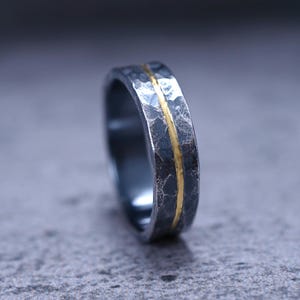 Puede incluir: Un anillo de metal gris oscuro con una superficie muy martillada y texturizada. Una banda delgada, lisa y pulida de color dorado recorre horizontalmente el centro del anillo, creando un contraste marcado con el acabado exterior oscuro y rugoso.