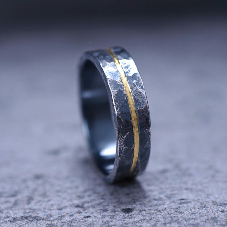 Puede incluir: Un anillo de metal gris oscuro con una superficie muy martillada y texturizada. Una banda delgada, lisa y pulida de color dorado recorre horizontalmente el centro del anillo, creando un contraste marcado con el acabado exterior oscuro y rugoso.