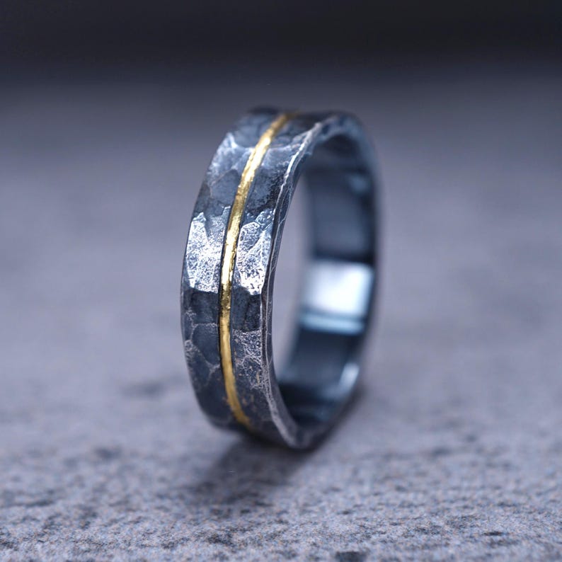 Puede incluir: Un anillo de metal martillado en color gris oscuro que presenta una banda delgada, lisa y de color dorado que recorre horizontalmente el centro de la superficie texturizada. El anillo tiene un perfil ancho y plano con un acabado rugoso.