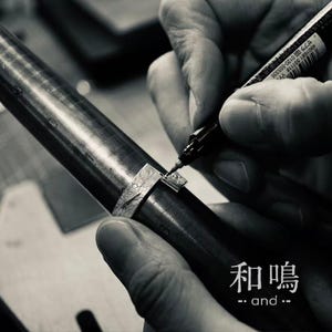 Puede incluir: Un anillo de plata con superficie texturizada se encuentra sobre un mandril de metal. Se utiliza un marcador negro para marcar el metal. El texto "和鳴 and" es visible en la esquina inferior derecha. La imagen es en blanco y negro.