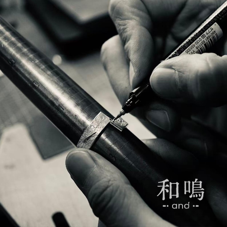Puede incluir: Un anillo de plata con superficie texturizada se encuentra sobre un mandril de metal. Se utiliza un marcador negro para marcar el metal. El texto "和鳴 and" es visible en la esquina inferior derecha. La imagen es en blanco y negro.
