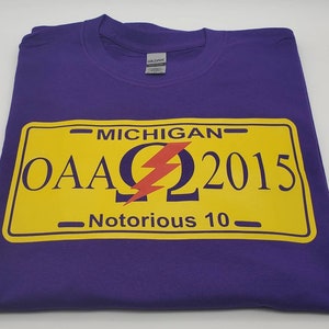 以下が含まれることがあります： 紫色のTシャツに黄色のナンバープレートのデザイン。ナンバープレートのグラフィックには、「MICHIGAN」の文字、「OAA」の文字、「2015」の数字、「Notorious 10」の文字が含まれています。中央には赤い稲妻があります。