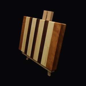 May include: A rectangular wooden cutting board with alternating stripes of light and dark brown wood. The board is supported by a small wooden easel, showcasing the butcher block design. The wood grain is visible.