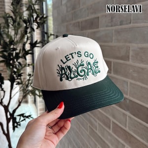 Könnte beinhalten: Eine cremefarbene Baseballkappe mit dunkelgrünem Schirm und dunkelgrünem Sticktext, der LET'S GO ALGAE lautet, mit stilisierten, algenartigen Buchstaben. Die Kappe hat eine strukturierte Krone und einen Knopf oben sowie den Markennamen NORSELAVI.