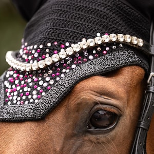 Op de afbeelding: Een zwart gehaakt oornetje voor paarden met een frontriem versierd met een rij heldere kristallen. Het stoffen paneel onder de frontriem is gedecoreerd met een dicht patroon van verspreide heldere en felroze strass-steentjes en een rand.