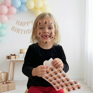 May include: A young person smiles, with stickers on their face and holding a sheet of stickers. The stickers are round with a brown and orange design. A banner that says "HAPPY BIRTHDAY" is in the background.