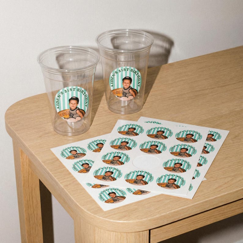 May include: Clear plastic cups and stickers with a birthday theme. The stickers and cups feature a photo of a young person and the text "AIDEN'S 21ST BIRTHDAY" in a circular design with a teal and white striped border.
