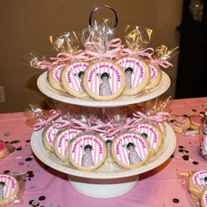 May include: A two-tiered white serving stand holds individually wrapped cookies. Each cookie has a pink and white striped border with a photo and the text "Sophia's 40th Birthday". The cookies are tied with pink ribbons.