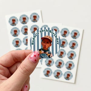 May include: A sheet of stickers featuring a photo of a person wearing a cap and overalls, with the text "Bodhi's 30th Birthday." The stickers have a blue and white striped background. Additional stickers with a similar design are visible.