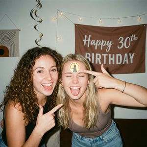 May include: Two smiling women celebrating a birthday. One woman has a sticker on her forehead that says "Always Beth Birthday". A brown banner in the background reads "Happy 30th Birthday". Decorative lights and a wall hanging are also visible.