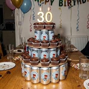 May include: A unique birthday cake made of stacked beverage cans, topped with chocolate cupcakes and a golden "30" candle. The cake sits on a wooden table, surrounded by party decorations like balloons and streamers.