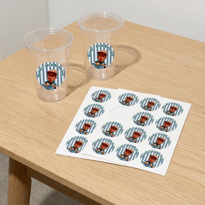 May include: Clear plastic cups and sticker sheets with a blue and white striped design. The stickers and cups feature a photo of a baby and the text "Bodhi's 30th Birthday" in a circular design. The items are on a light wood surface.