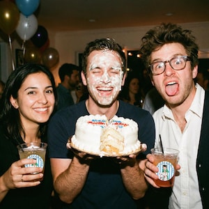 May include: Three people smiling at a birthday party. One person has cake frosting on their face and is holding a white cake with the words "Happy Birthday" on it. The other two people are holding drinks. Balloons are in the background.
