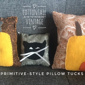 May include: Three handmade felt cushions with primitive-style designs. The cushions feature a black cat, a yellow pumpkin, and a yellow pumpkin with an orange stem. The cushions are arranged on a grey sofa with the text "COTTONTAIL VINTAGE" and "PRIMITIVE-STYLE PILLOW TUCKS" visible.