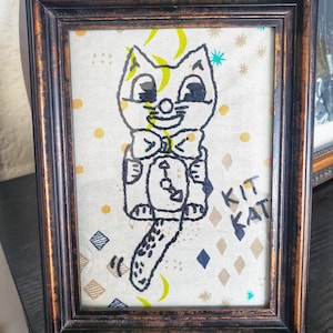 May include: A framed embroidery of a cartoon cat wearing a bow tie and holding a clock. The embroidery is on a white fabric with a light brown background and the words "Kit Kat" are stitched in the bottom right corner.