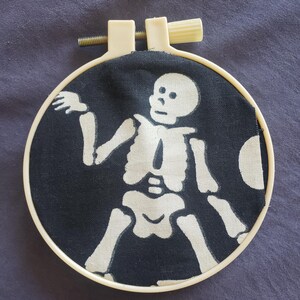 May include: A black fabric with a white skeleton design stretched within a cream-colored embroidery hoop. The skeleton is depicted with outstretched arms and legs. The hoop is secured with a screw and a tightening knob.