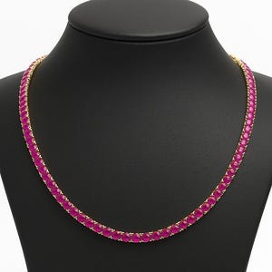 May include: A gold necklace featuring a continuous line of magenta gemstones. The necklace is displayed on a black jewellery bust against a white background, emphasising the piece's colour and design.