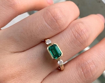 May include: A gold ring featuring a rectangular, emerald-cut green gemstone, flanked by two smaller, round, clear gemstones. The ring is set in a gold band, showcasing a classic and elegant design.
